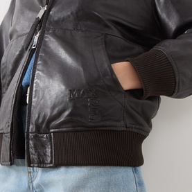 Model wearing Max&Co dark brown leather bomber jacket, close-up of ribbed hem, zip detail and embossed logo, styled with light blue jeans.