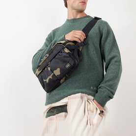Model carrying Porter Yoshida Camouflage Belt Bag in woodland khaki, worn crossbody, with green knit sweater and cream trousers