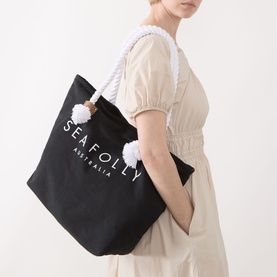 Model carrying Seafolly Ship Sail Tote Bag in black canvas with white rope handles, pom-pom knot detail and contrast logo branding.