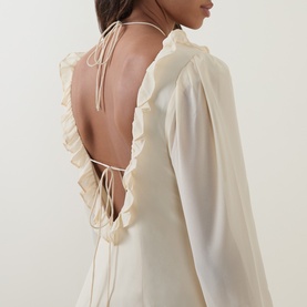 Model wearing Anne Louise Boutique cream dress with open ruffle-trimmed back and tie-neck detail, long sleeves visible