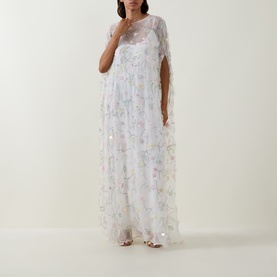 Model wearing Bernadette dress in Clematis Emb White, sheer floral embroidered maxi with cape sleeves and sequin detailing, styled with heeled sandals