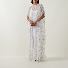 Model wearing Bernadette dress in Clematis Emb White, floor-length sheer floral overlay with cape sleeves and slip lining beneath.