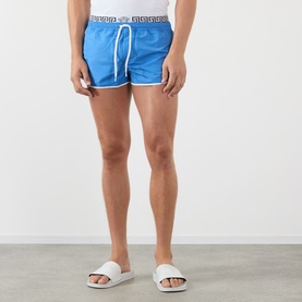 Model wearing Versace azure swim shorts with Greca logo waistband and white drawstring, styled with white slides