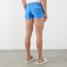 Model wearing Versace azure swim shorts, rear view, with white trim detail and branded waistband, styled with white slider sandals.