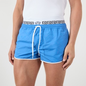 Model wearing Versace azure swim shorts with white trim, Greek key Greca waistband, and white drawstring, styled with a white vest.
