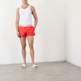 Model wearing Versace vintage red swim shorts with logo waistband, styled with a white vest and white slides.