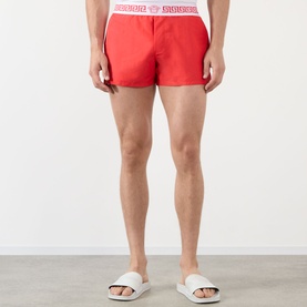 Model wearing Versace vintage red swim shorts with branded white elasticated waistband, styled with white slides.