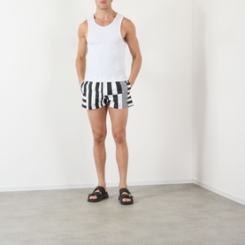 Model wearing Versace black and white bold stripe swim shorts with white vest top and black slider sandals.