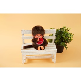 Monchhichi plush soft toy in dark brown with red bib, seated on a miniature white wooden bench