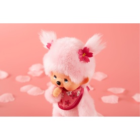 Monchhichi pink plush toy with floral hair accessories, cherry blossom details, and a pink bib, photographed against a soft pink background.