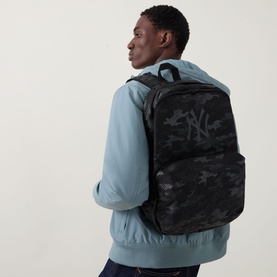 Model carrying New Era Logo Camo Backpack in black camo print with tonal NY logo, adjustable straps, styled with light blue jacket