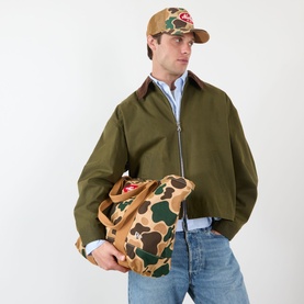 Model carrying New Era Duck Camo Tote Bag in multi camo print with grab handles, styled with olive jacket and matching camo cap