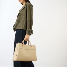 Model carrying Mario Valentino Alexia Tote Bag in beige with gold-tone hardware and top handles, styled with olive jacket and navy trousers