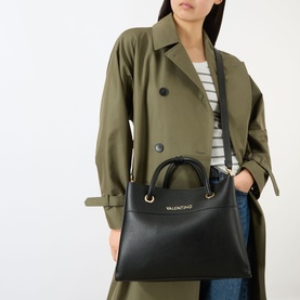 Model carrying Mario Valentino Alexia Logo Tote Bag in black with gold-tone hardware and top handles, styled with olive trench coat