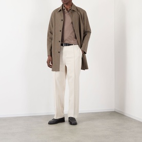 Model wearing a taupe single-breasted mac coat with cream tailored trousers, brown polo shirt, and black leather loafers
