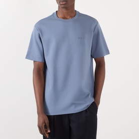 Model wearing blue crew neck short sleeve t-shirt with small chest logo, styled with black trousers
