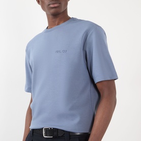 Model wearing pale blue oversized crew neck T-shirt with tonal chest logo, styled with black belted trousers