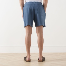 Model wearing Paige Elijah Hidden Drawstring Shorts in Lakeside blue, rear view, with brown sandals visible.