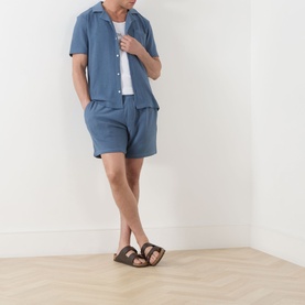 Model wearing Paige Elijah Hidden Drawstring Shorts in Lakeside blue, styled with a matching blue camp-collar shirt, white tee and brown sandals.