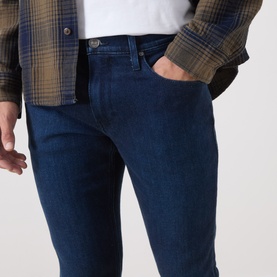 Model wearing Paige 3049 Straight Jeans in Pembry dark blue denim, styled with a white tee and olive plaid overshirt.