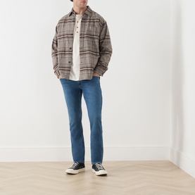 Model wearing Paige Bushman straight jeans in mid-blue, styled with a brown plaid overshirt, white tee and black high-top trainers.