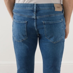 Model wearing Paige Straight Jeans in Bushman mid-blue denim, rear view showing back pockets and slim straight leg.