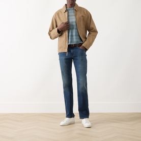 Model wearing Paige Kirkwin straight jeans in mid-blue wash, styled with a tan jacket, striped polo and white trainers.