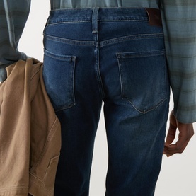 Model wearing Paige Kirkwin straight jeans in dark indigo wash, rear view, styled with a blue check shirt and tan jacket.