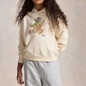 Model wearing Ralph Lauren Patchwork Polo Bear Logo Terry Hoodie in Guide Cream with patchwork bear appliqu&eacute;, styled with stripe trousers.