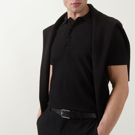 Model wearing Tom Ford black cotton polo shirt with three-button placket, styled with black trousers and belt, jacket draped over shoulder.