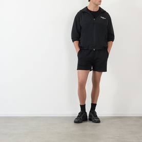 Model wearing 247 Jet Black cotton shorts with matching zip-up jacket and black trainers, hands in pockets.