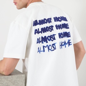 Model wearing Almost Home T-Shirt in white with repeated blue printed logo text across the back, oversized fit