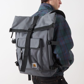 Model carrying Carhartt Philis Logo Backpack in Styx grey, roll-top flap with black straps and buckles, front zip pocket, worn over plaid jacket