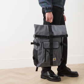 Model carrying Carhartt Philis Logo Backpack in Styx grey, held by top handle, with black roll-top flap, front pockets and black hardware.