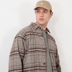 Model with Carhartt Madison Logo Cap in Leather/White, six-panel unstructured cotton cap with embroidered logo, styled with brown plaid overshirt