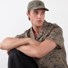 Model with Carhartt Travon Duck Feather Cap in duck feather print olive and black, flat peak, styled with matching printed camp collar shirt