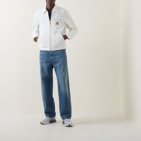 Model wearing Carhartt Detroit Casual Jacket in Wax white with tonal corduroy collar and front zip, styled with blue jeans and grey trainers.