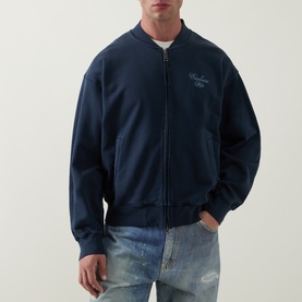 Model wearing Carhartt casual jacket in Carbon navy, zip-front bomber style with embroidered chest script, styled with light wash distressed jeans