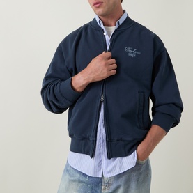 Model wearing Carhartt Casual Jacket in Carbon navy, zip-front with script logo, styled with blue striped shirt and light-wash jeans.