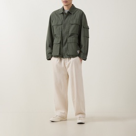 Model wearing Carhartt Mandelson Logo Field Jacket in Leaf green with multiple patch pockets, styled with cream wide-leg trousers and white trainers.