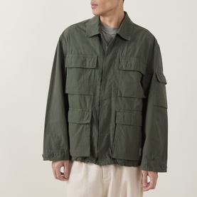 Model wearing Carhartt Mandelson Logo Field Jacket in Leaf green, styled with cream trousers, featuring multiple flap patch pockets and flat collar.