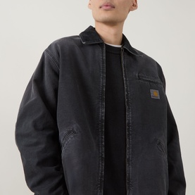 Model wearing Carhartt OG Detroit Jacket in Black, showing zip fastening, chest pocket, and woven logo badge, styled with a black crew-neck top.