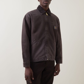 Model wearing Carhartt OG Detroit Logo Jacket in Tobacco. Dark brown canvas, shirt collar, front zip, logo badge. Styled with black trousers.