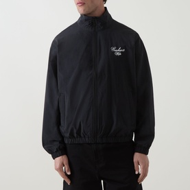 Model wearing Carhartt Travon Logo Windbreaker in black, showing stand collar, front zip, elasticated trims and chest logo embroidery.