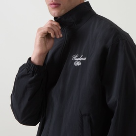 Model wearing Carhartt Travon Logo Windbreaker in black, showing chest logo embroidery and stand collar with front zip fastening.