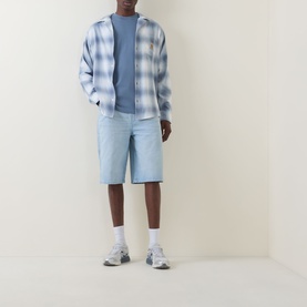 Model wearing Carhartt Tyers Logo Check Print Shirt in Shady Blue check, styled with light blue shorts and grey trainers.