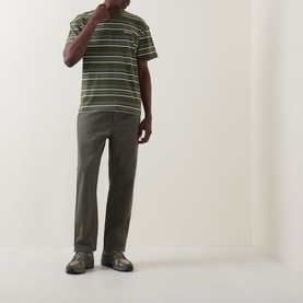 Model wearing Carhartt Flint Casual Trousers in Leaf, tapered-leg fit, styled with a green striped tee and dark chunky sandals.