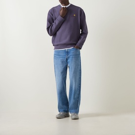 Model wearing Carhartt American Script Logo Sweatshirt in Shady Purple, crew neck with chest logo, styled with blue wide-leg jeans and grey trainers.