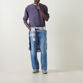 Model wearing Carhartt American Script Logo Sweatshirt in Shady Purple with blue jeans and a shirt tied at the waist, grey trainers.