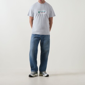 Model wearing Carhartt Bumper Stickers Logo T-Shirt in white with blue straight-leg jeans and grey trainers.